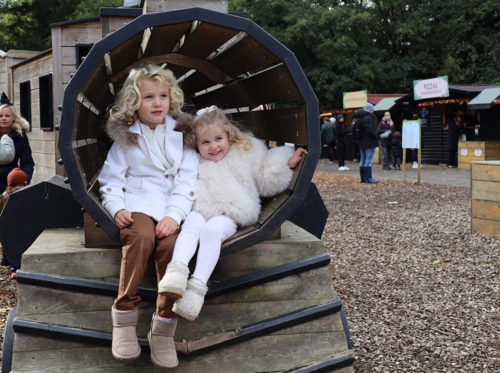 Two children playing on wooden train in playground surrounded by trees.