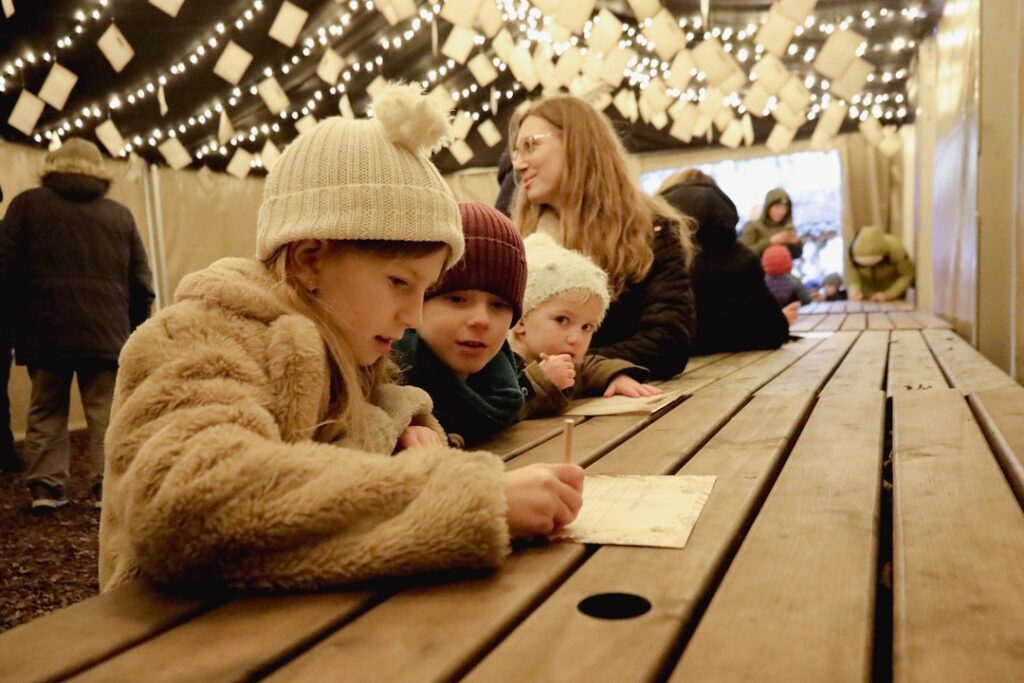 Young children writing letters to Father Christmas in a festive wooden lodge decorated with twinkling fairy lights at Audley End Enchanted Railway’s Christmas Special in Essex.