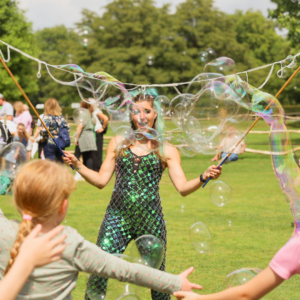 Performer with teal hair blowing large bubbles while children reach out in a sunny park.