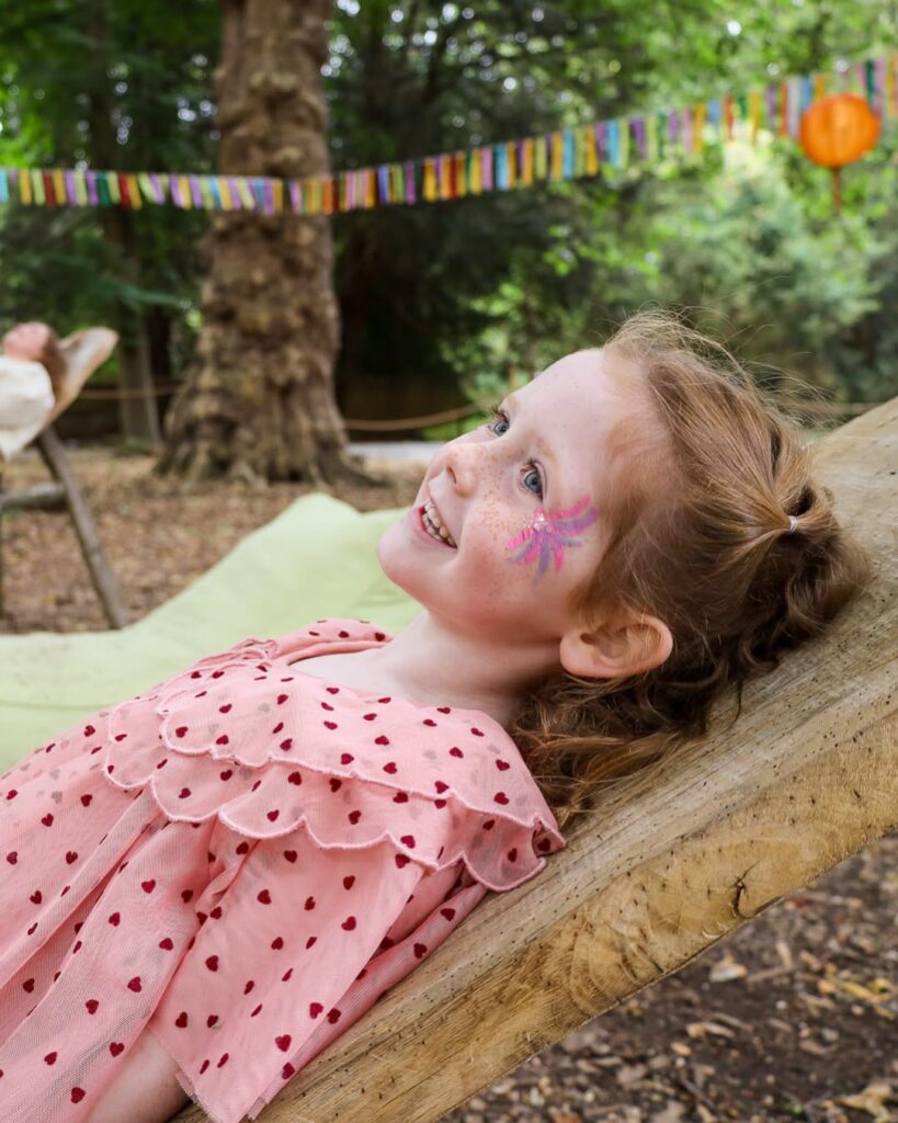 Child relaxing in a woodland hammock with fairy face paint at Audley End Enchanted Railway's Summer Festival.
