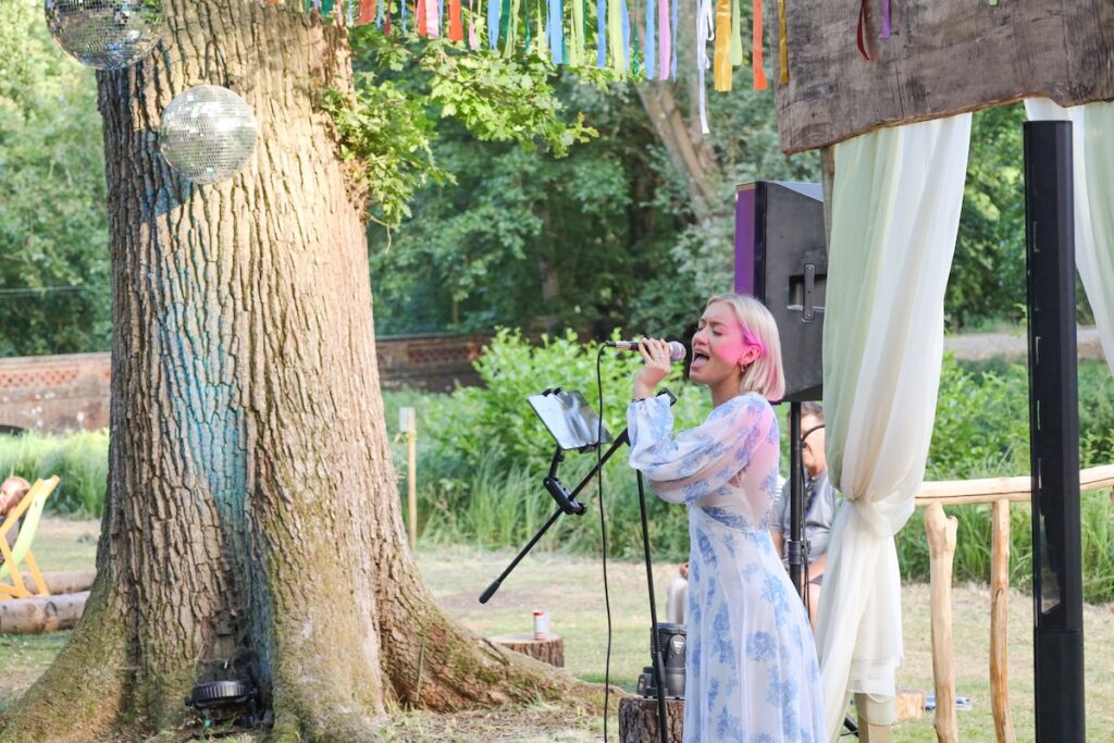 Female singer with performing outdoors at Rivernights under a tree, singing into a microphone with a stand, colorful ribbons and disco balls overhead.