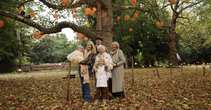 Family of six posing under a tree with hanging jack-o'-lanterns in a leaf-strewn park (autumn/Halloween).