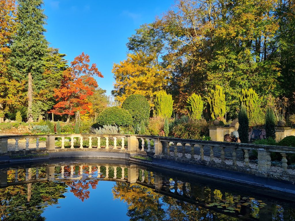 Autumn trees and clipped shrubs beside a garden pond, with a stone balustrade reflected in the water.