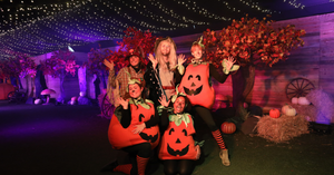 Group of six people in pumpkin costumes posing and waving at a autumn-themed Halloween display.