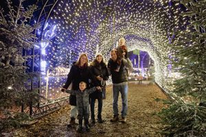 Family posing together under a tunnel of warm holiday lights in a snowy outdoor display, smiling at the camera.