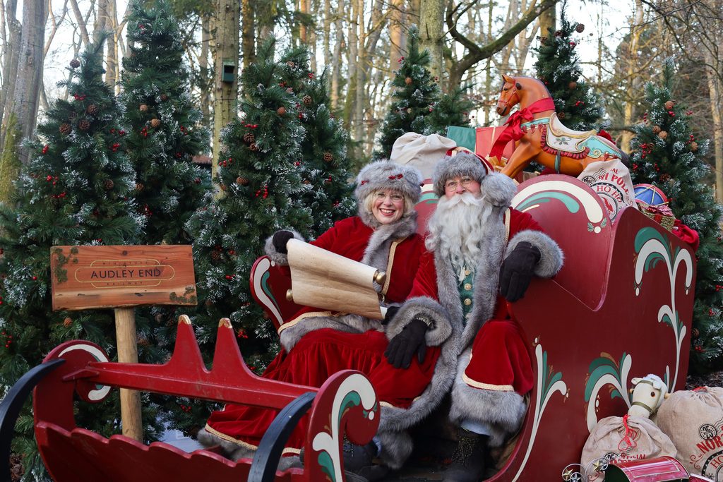 Santa Claus and Mrs. Claus in red costumes ride a decorative red sleigh float among snow-dusted evergreen trees, at Audley End Railway.