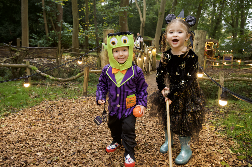 Two kids in Halloween costumes walk along a leaf-covered path in a wooded area, one in a green Frankenstein mask and purple suit, the other in a black sparkly witch costume with ears, lanterns glowing in the background.