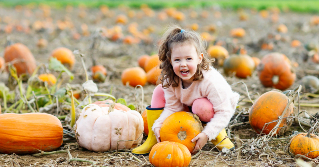 A smiling young girl in yellow wellies crouches in a pumpkin patch, holding an orange pumpkin.