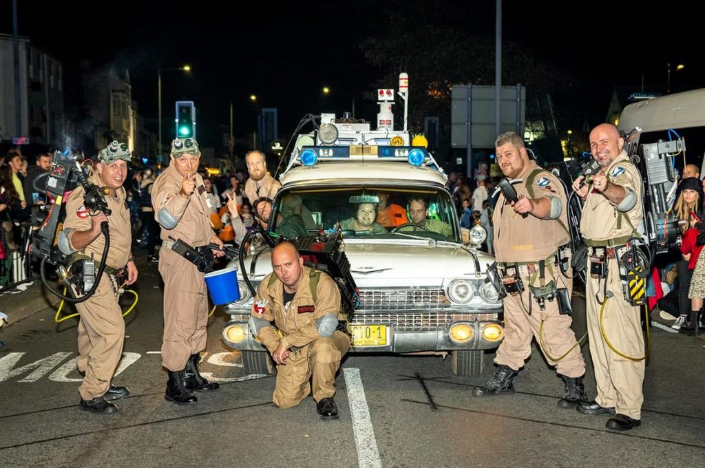A group dressed as Ghostbusters pose around a replica Ecto-1 car on a crowded street at night.