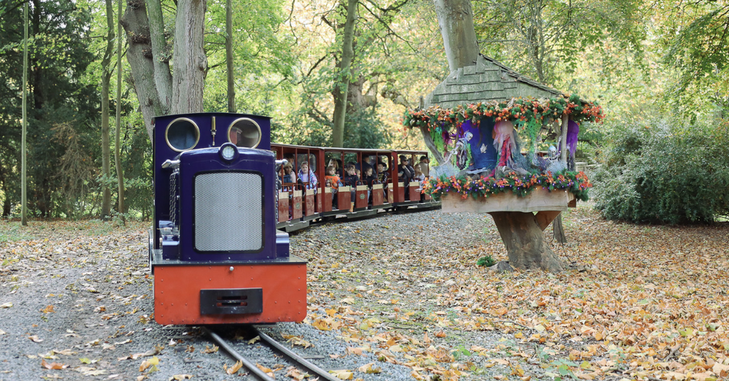 Blue miniature train carrying families through autumn woodland at Audley End Enchanted Railway’s Halloween Special, passing a colourful, Halloween-themed fairy treehouse.