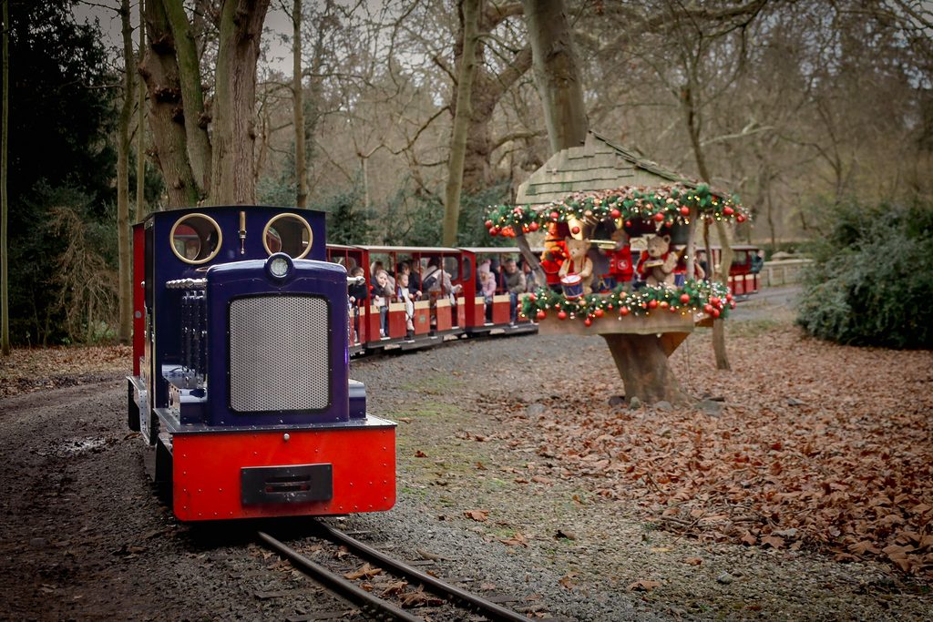 Small vintage steam train pulling red carriages with passengers along a leaf-strewn park path, autumn trees in the background and a festively decorated float nearby.