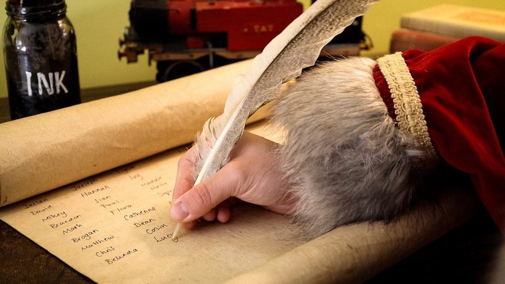 Close-up of Father Christmas writing names on a parchment scroll with a feather quill at Audley End Enchanted Railway’s Christmas Special in Essex.