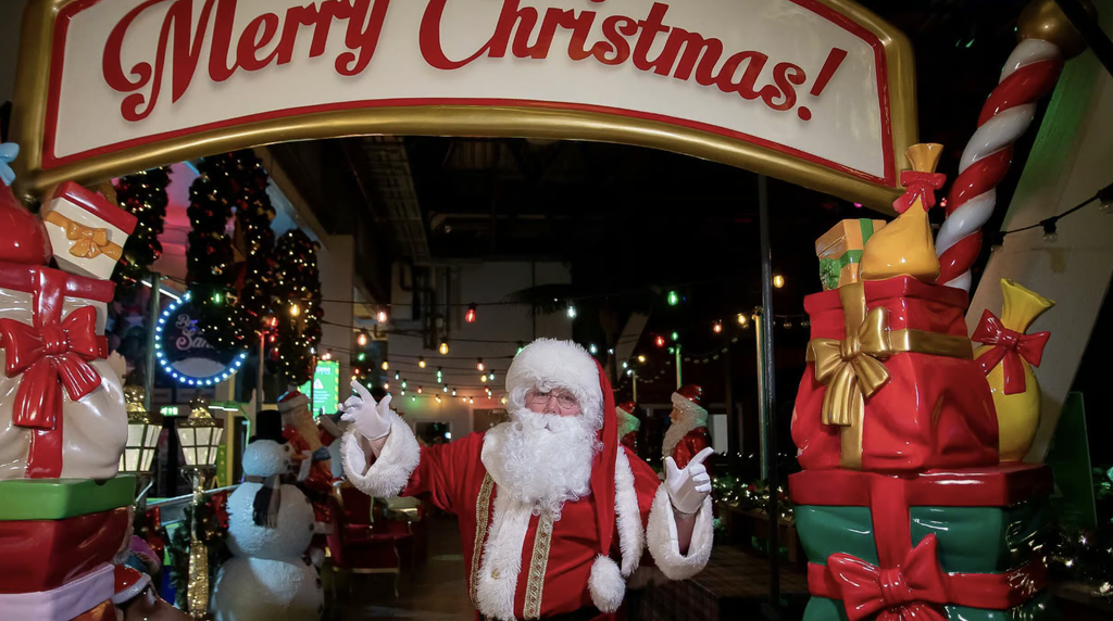 Father Christmas waving beneath a large Merry Christmas archway, surrounded by colourful presents, festive decorations and twinkling lights at a magical Christmas attraction.