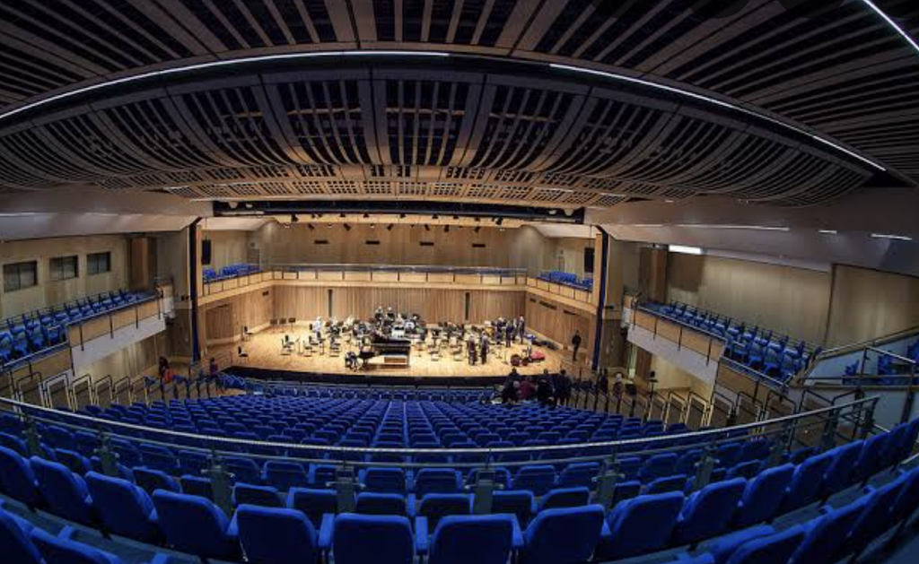 Interior of a modern concert hall with rows of blue seats, balcony seating and a large wooden stage set up for an orchestra performance.