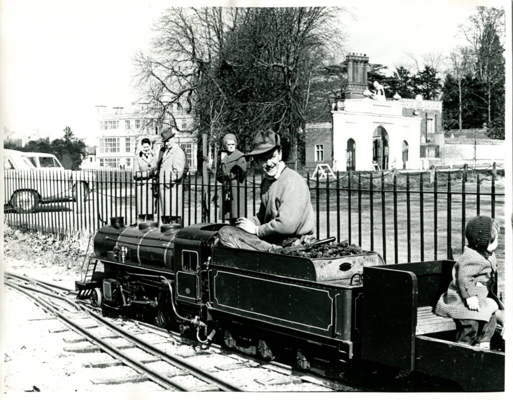 Historic black-and-white photograph of a miniature steam locomotive at Audley End Railway in Essex, with a driver and young passenger aboard and Audley End House visible in the background.