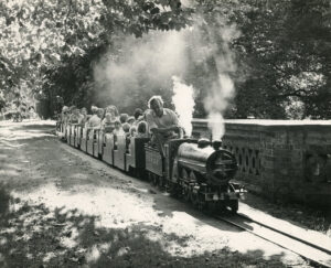 Historic black-and-white photograph of families riding a miniature steam train at Audley End Railway in Essex, with steam rising from the locomotive as it travels through the woodland.